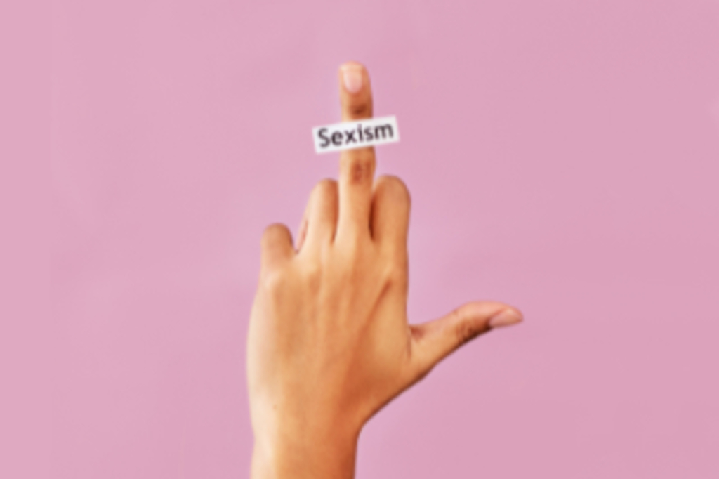 Studioaufnahme einer nicht erkennbaren weiblichen Hand, die den Mittelfinger zeigt. Auf dem Finger klebt ein Zettel mit dem Wort ‚Sexismus'. Der Hintergrund ist rosa., Link auf Frauenbilder I Männerbilder I Körperbilder - Stadt Köln
