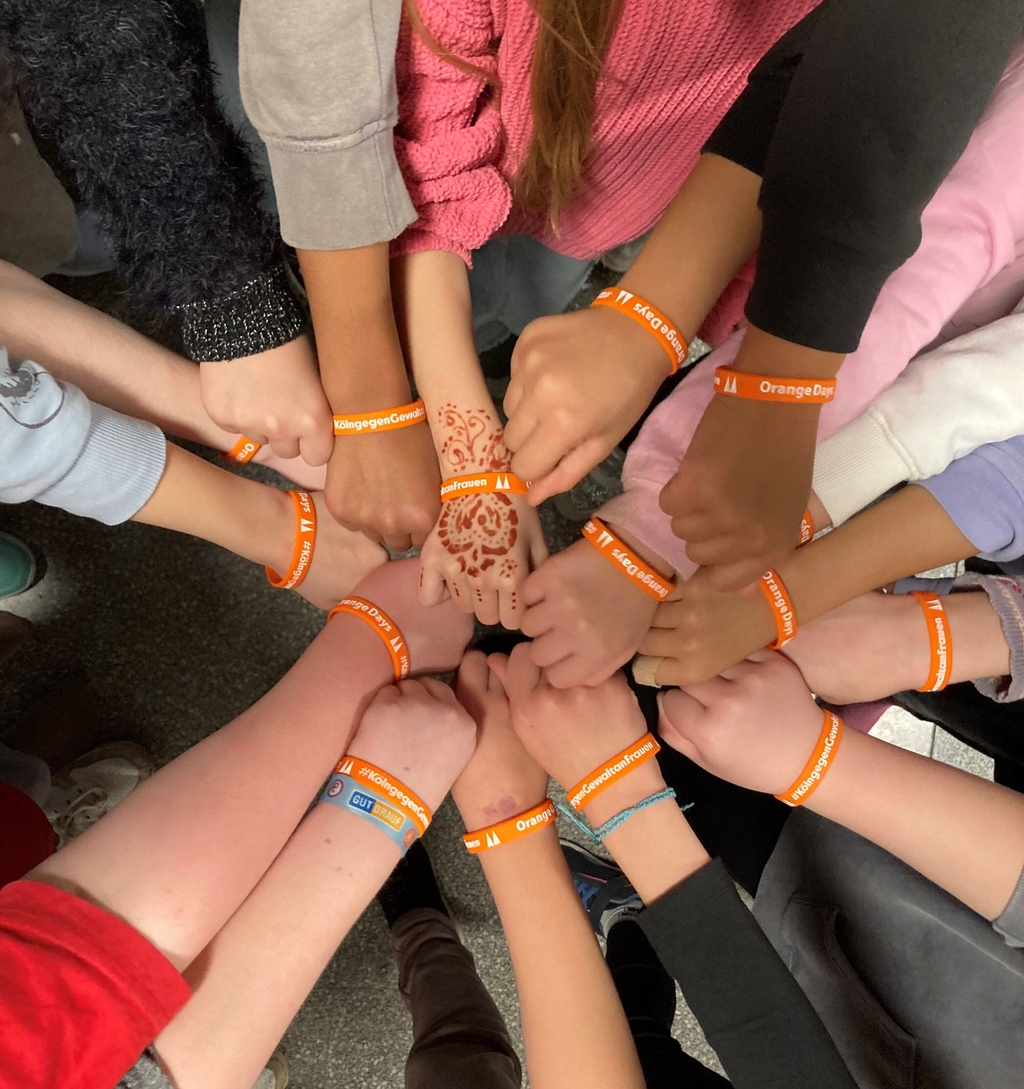 Mehrere Kinder stehen in einem Kreis und legen ihre Hände in die Mitte. Alle tragen orangefarbene Armbänder mit der Aufschrift Orange Days