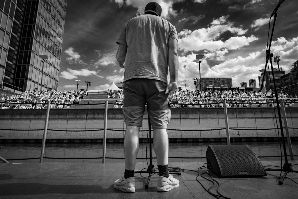 Bild von einem Slam-Poeten von hinten. Im Hintergrund sitzen die Zuschauer*innen auf der Bühne