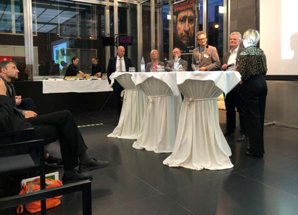 Stehtische, an denen Personen stehen und die Diskussion führen. Ein Besucher auf einem Stuhl ist zu erkennen.