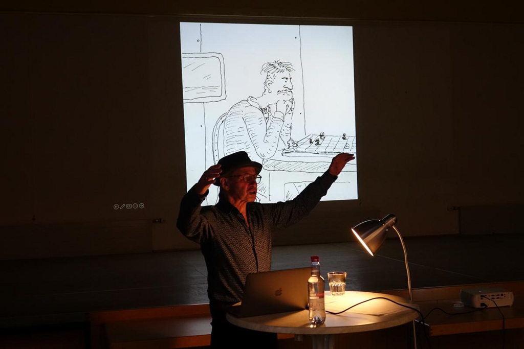 Didi Jünemann steht gestikulierend vor Leinwand mit Comic, der einen Schachspieler zeigt