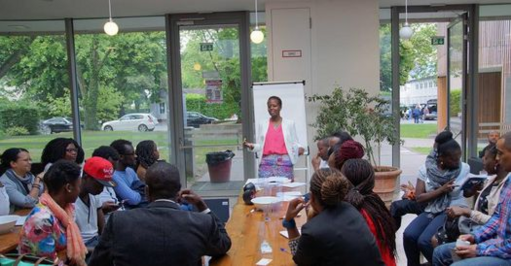 Frau (PoC) steht erzählend vor Flipchart, weitere Personen sitzen davor und hören zu 