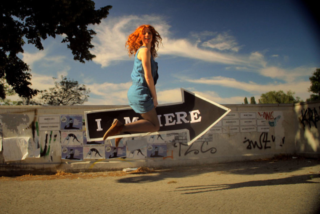 Rothaarige Frau in blauem Kleid sitzt lachend auf einem Pfeil und schau in die Kamera. Der Pfeil trägt den Schriftzug " I am here"