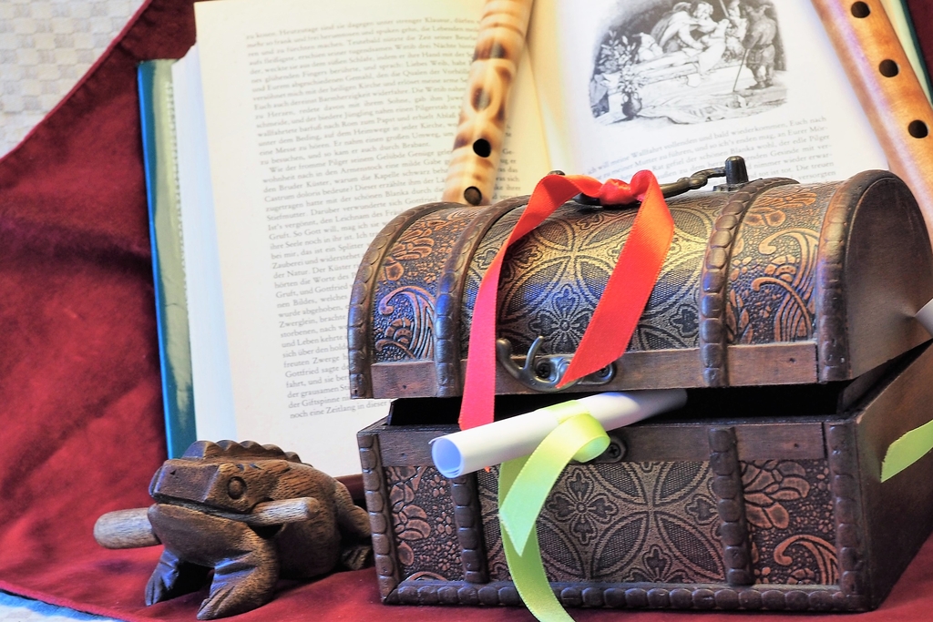 Kleine, braune Kiste mit Schleifen in Rot und Grün, darin eine kleine Papierrolle; daneben ein aus Holz geschnitzter Frosch mit Knüppel im Mund. Dahinter ein aufgeschlagenes Buch und zwei Blockflöten aus Holz. 