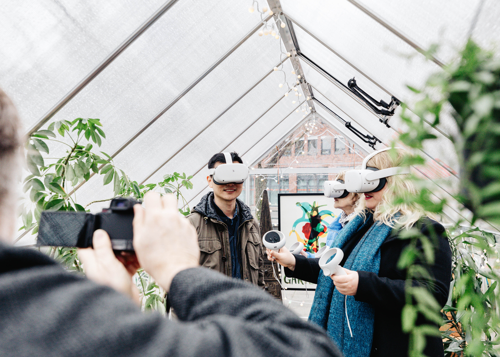 Vier Personen in einer Art Gewächshaus. Drei davon haben eine VR-Brille auf dem Kopf, die vierte Person filmt.