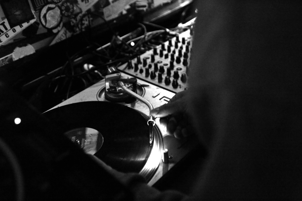 Foto eines Plattenspielers und Mischpults in schwarz-weiß. Eine Person (nur teilweise abgebildet) hält seinen Daumen auf den Plattenspielerarm.