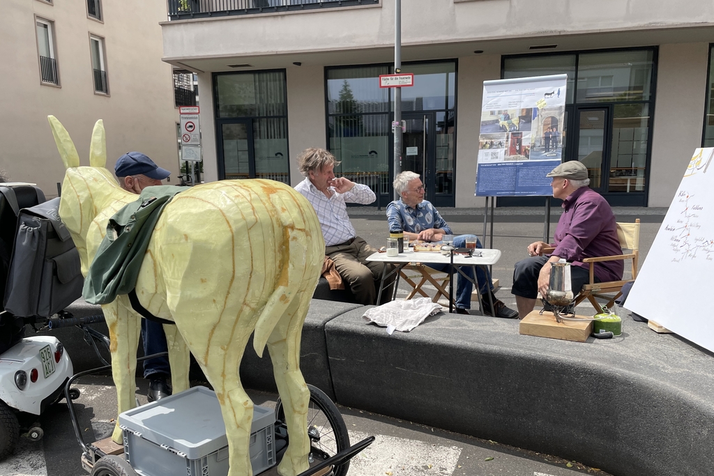 Eine Eselsklulptur im Vordergrund, im Hintergrund drei Männder, die an einem Tisch sitzen und sich unterhalten