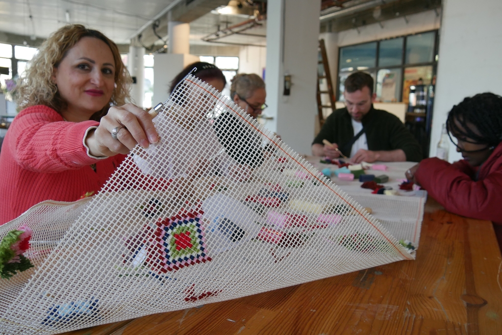 Mehrere Personen sitzen zusammen und basteln. Eine Frau im vorderen Bildbereich zeigt ein gesticktes Motiv in die Kamera. 