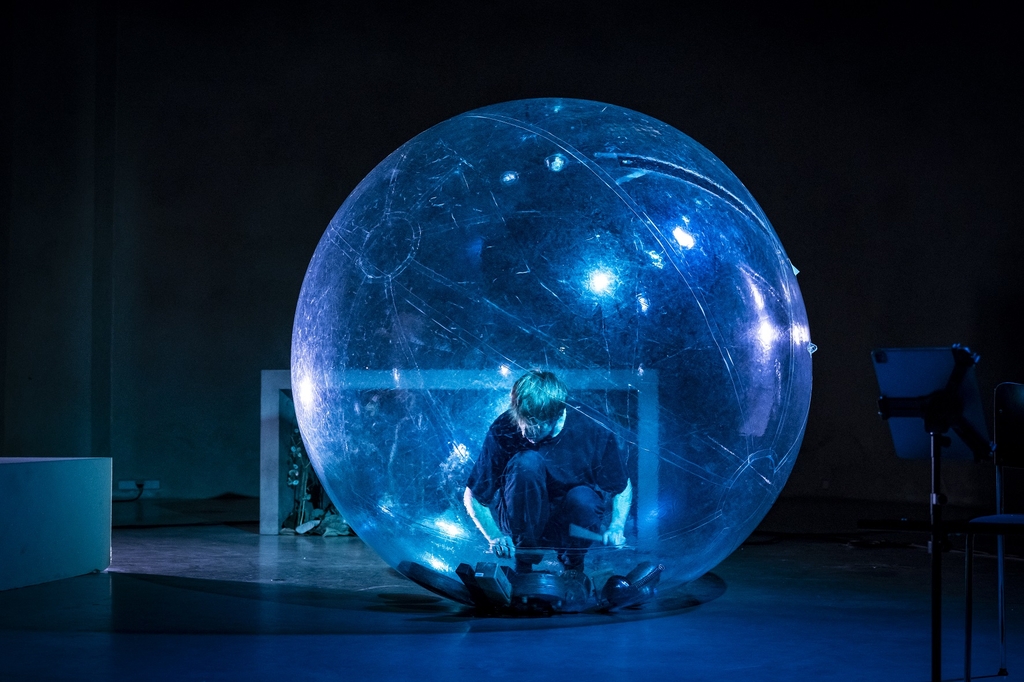 Eine große blau angeleuchtete Plastikkugel, in der ein*e Künstler*in sitzt und Musik macht