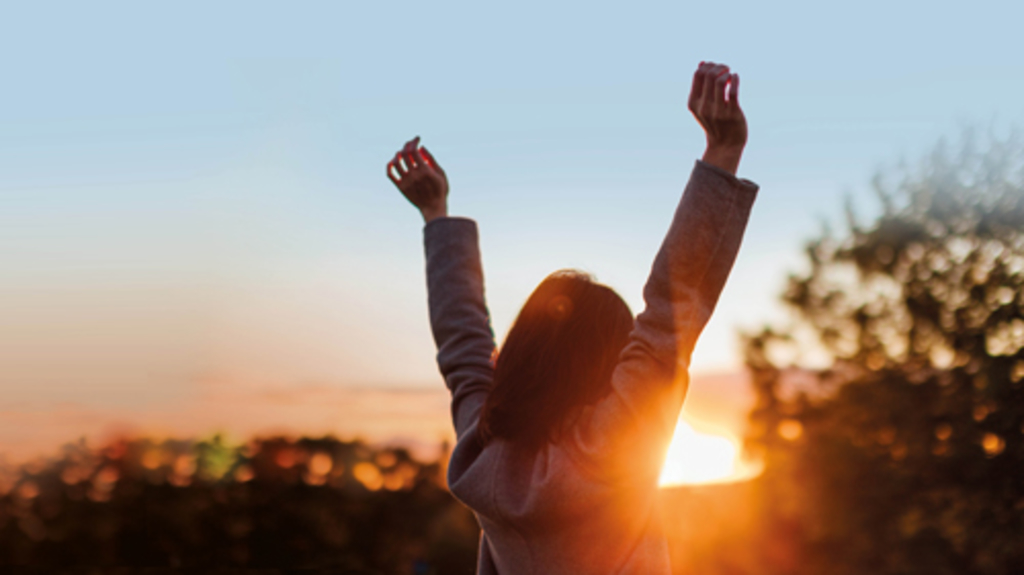 Sonnenuntergang, davor eine Frau von hinten fotografiert, reißt die Arme hoch