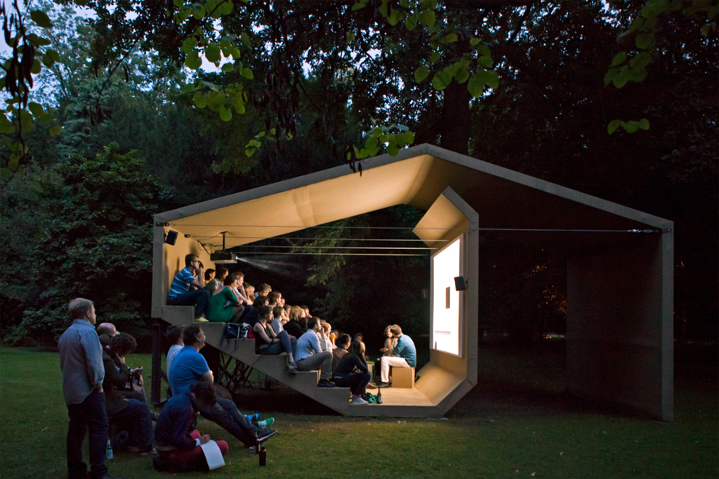 Menschen sitzen in einer Holzkonstruktion und schauen auf eine integrierte Leinwand; die Holzkonstruktion steht im Grünen, Link auf Open Air - Stadt Köln