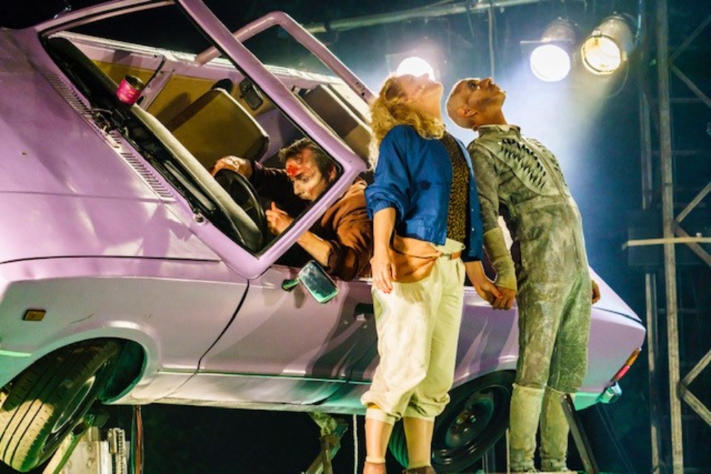 Zwei Darsteller*innen auf der Bühne die nach oben schauen, dahinter ein Mann, der in einem lilanen Auto hängt