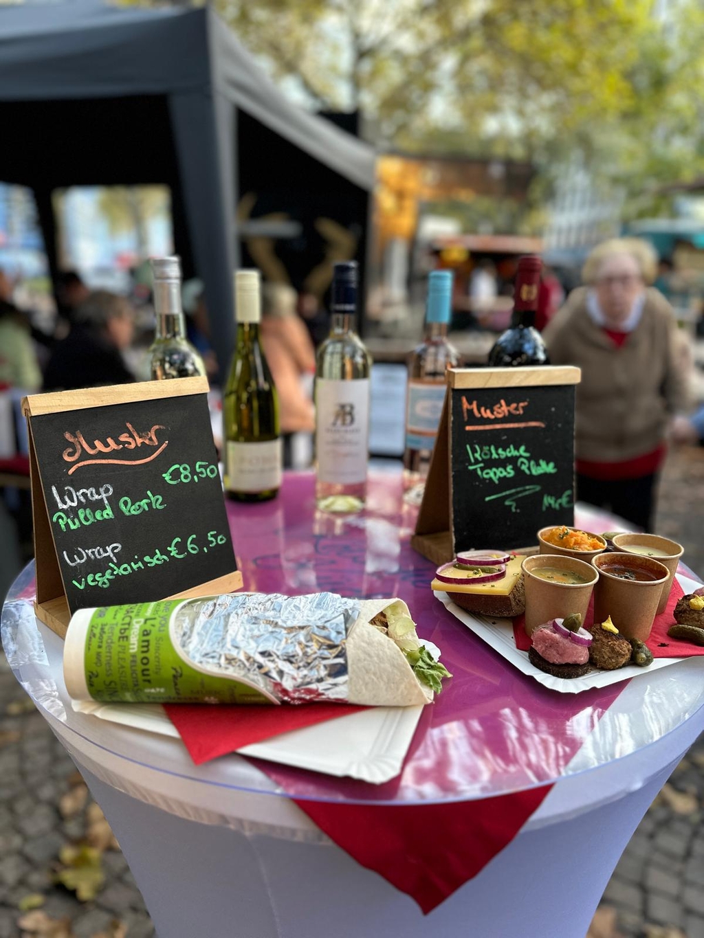Ein Wrap liegt auf einem Stehtisch, rechts daneben weitere Speisen. Im Hintergrund Besucher des Wochenmarktes.