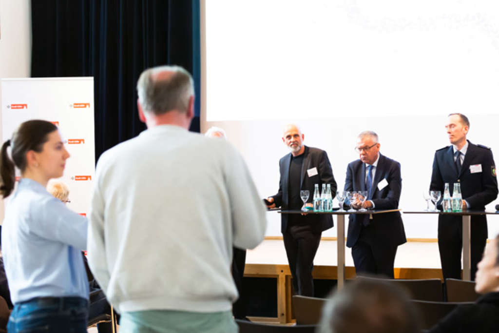 Bürger von hinten, der eine Wortmeldung macht, beim Stadtgespräch, vorne am Podium stehen Moderator Marc Jaschik, Amtsleiter Josef Ludwig (Amt für Wohnungswesen) und Gregor Eisenmann (Leiter der Kölner Polizeiinspektion 1)