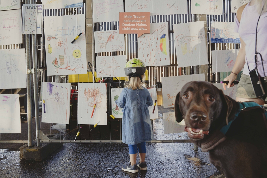 Zu sehen ist ein Bauzaun, an dem eine Sprechblase mit der Aufschrift „Mein Traumspielplatz am Deutzer Hafen sieht aus, wie…“, an dem viele Kinderzeichnungen aufgehängt sind am Hafentag. Davor steht ein kleines Mädchen und ein Hund ist zu sehen. 