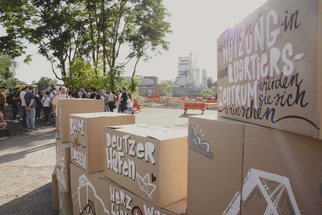 Zu sehen ist der Essiggarten in Köln am Hafentag. Hier sind mehrere Kartons bemalte und beschriftete Kartons aufgebaut, auf denen steht: „Welche Nutzung in einem Quartierszentrum würden Sie sich wünschen?“. Im Hintergrund sieht man eine größere Gruppe Menschen. 