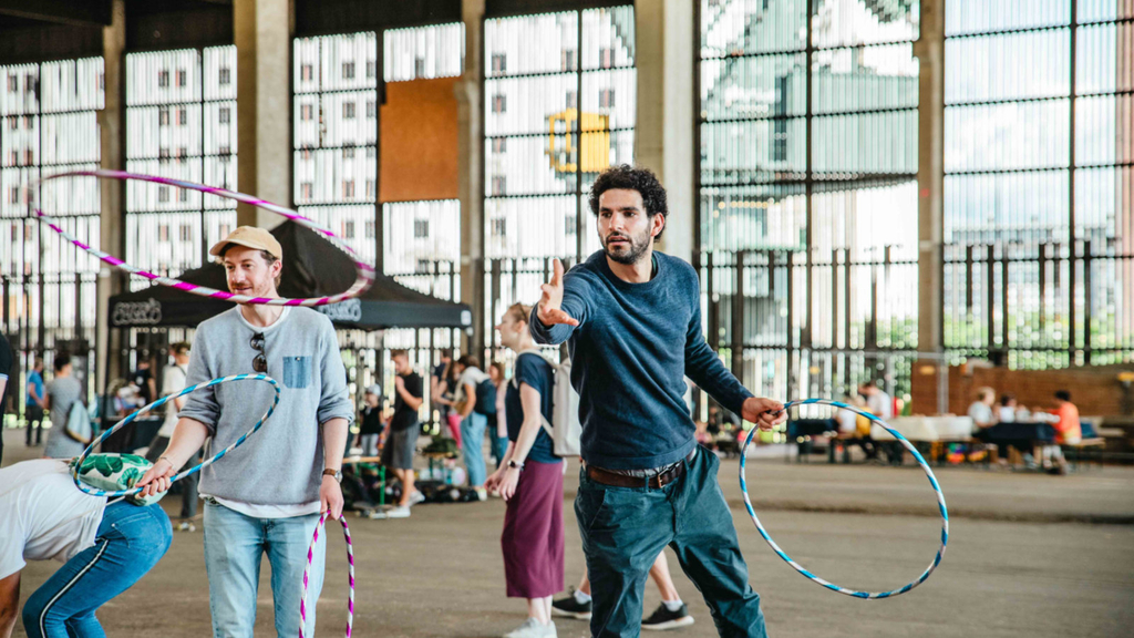 In der Holzhalle werfen zwei Personen bunte große Ringe auf Kegel, im Hintergrund sitzen Menschen an Bierbänken.