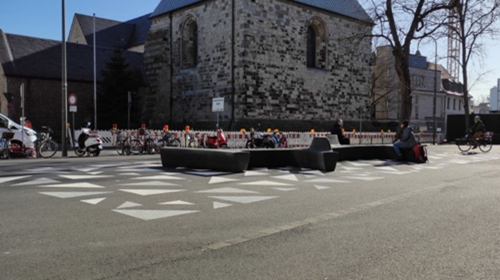Der Waidmarkt in der Kölner Innenstadt mit Blickrichtung nach Südosten auf St. Georg. Auf der Platzfläche sind verschieden große Dreiecke in den Farben Weiß und Hellgrau aufgemalt. Darauf stehen Sitzbänke, auf denen Personen sitzen. Rechts im Bild ist eine Person auf einem Fahrrad. Hinter der Platzfläche sind Baustellen-Zäune und abgestellte Zweiräder zu sehen.
