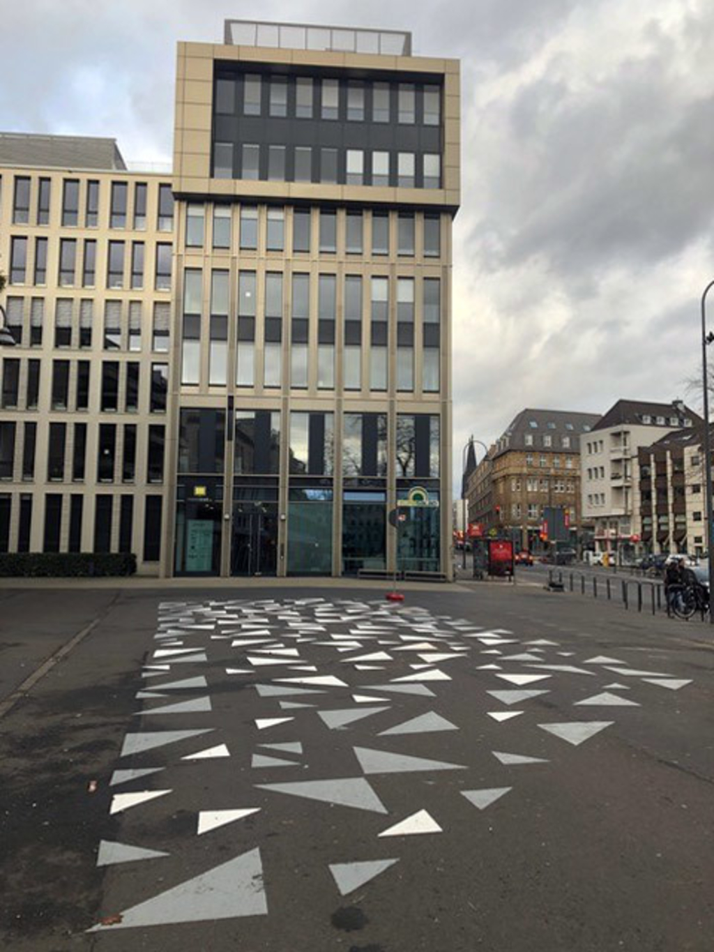 Das Bild ist ein Foto vom Waidmarkt in der Kölner Innenstadt mit Blickrichtung nach Norden. Darauf ist die Bebauung im Umfeld zu sehen. Im Fokus steht ein siebengeschossiges Gebäude. Vor diesem Gebäude ist eine größere Platzfläche zu sehen. Auf dem Asphalt dieser Platzfläche sind verschieden große Dreiecke in den Farben Weiß und Hellgrau aufgemalt.