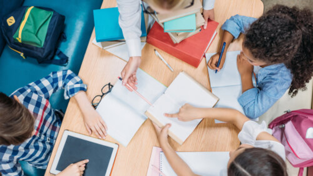 Blick von oben auf Lehrekraft und jungen Schüler*innen, die gemeinsam Hausaufgaben machen, Link auf Anmeldung an einer weiterführenden Schule - Stadt Köln