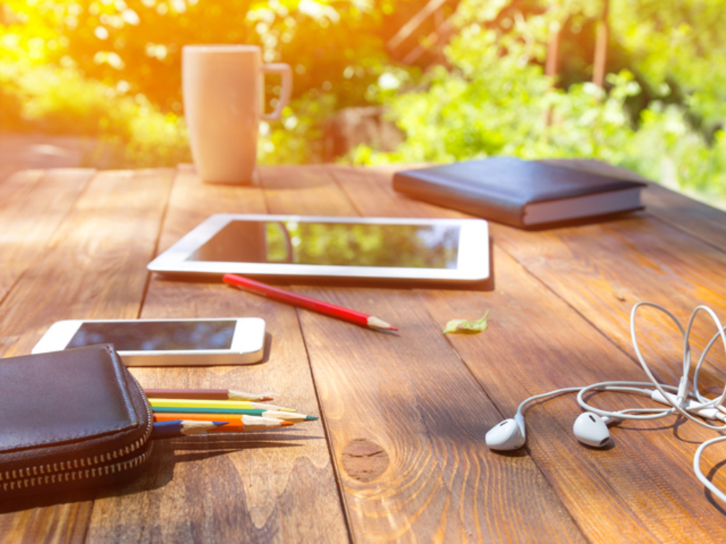 Hölzerner Schreibtisch auf dem Buntstifte, ein Smartphone, ein Tablet, Kopfhörer und ein Notizbuch liegen. Im Hintergrund steht eine Tasse.