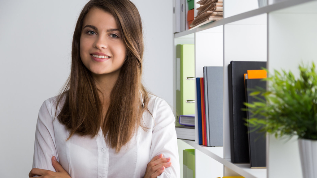 Eine junge lächelnde Geschäftsfrau steht mit verschränkten Armen im Büro neben einem Bücherregal und blickt in die Kamera.