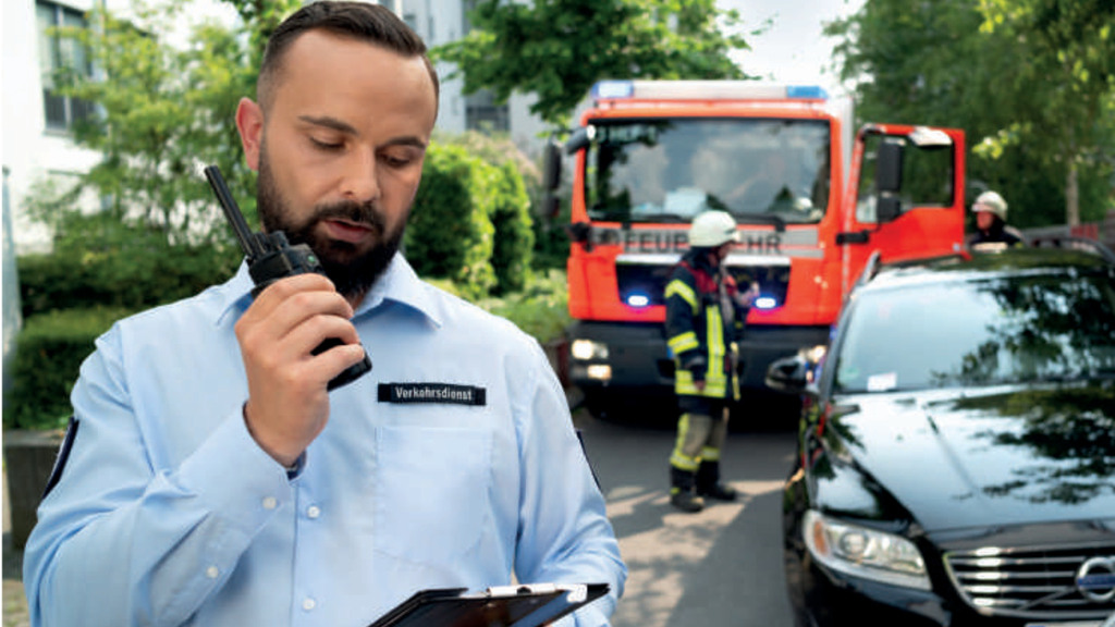 Mitarbeiter vom Verkehrsdienst spricht in ein Funkgerät, dahinter ein falschparkendes Auto, das ein Rettungsfahrzeug behindert., Link auf Verkehrsdienst - Verkehrsüberwachung - Stadt Köln