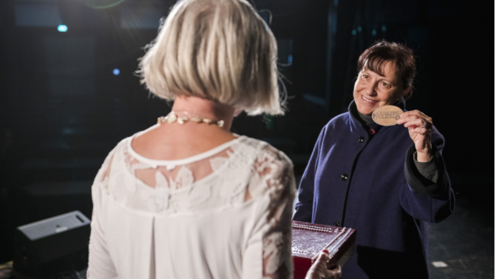 Eine ältere Dame von hinten, die eine Kiste hält, von vorne eine lächelnde Frau mit einem Emblem, auf dem Kriminalppolizei steht.