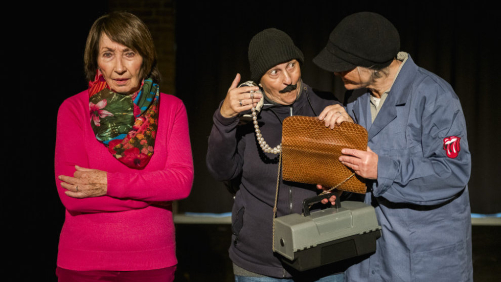 Auf einer Bühne stehen links eine besorgte ältere Dame, rechts 2 Handwerker, die in eine Handtasche schauen