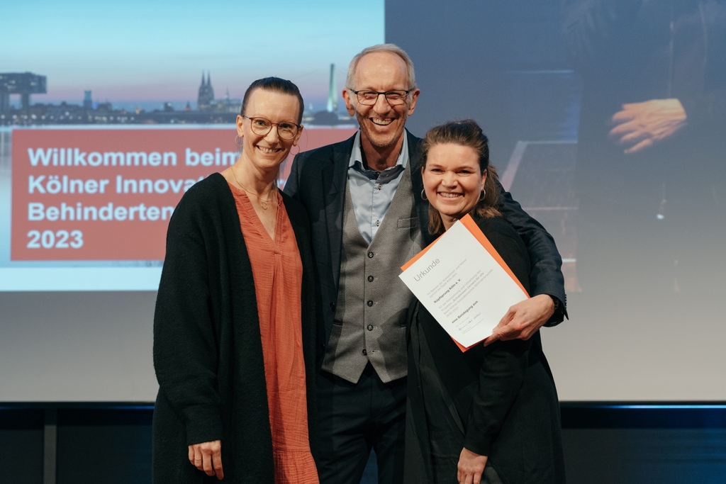 Drei Vertreter*innen von Kopfsprung e. V. stehen auf einer Bühne und halten eine Urkunde in der Hand. 