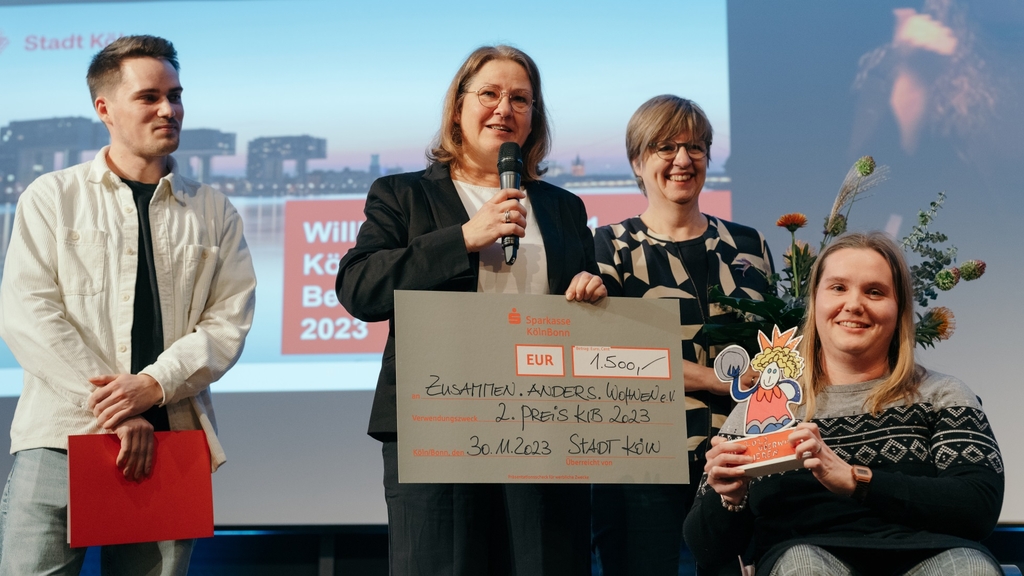 Vertreter*innen von Zusammen. Anders. Wohnen. e. V. halten Preis und Scheck in der Hand. Eine Frau hält ein Mikrofon in der Hand.