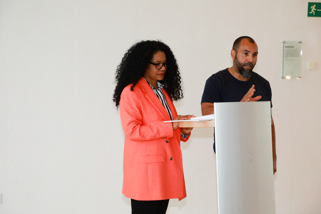 Zwei Mitglieder des Expert*innengremiums stehen am Pult und sprechen zum Publikum. Die Frau steht links und trägt eine korallfarbene Jacke, der Mann steht links und trägt ein schwarzes T-Shirt. 