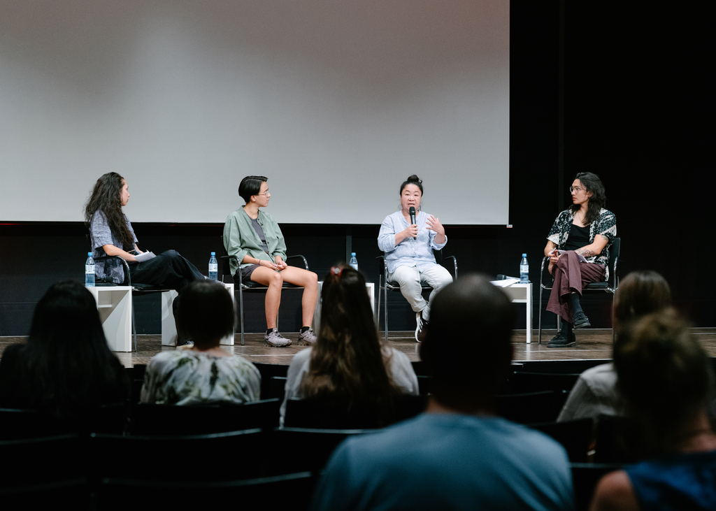 Vier Menschen sitzen auf Stühlen auf einer Bühne und diskutieren. Von hinten sind Zuschauer*innen zu sehen, die den Kopf auf die Bühne richten.