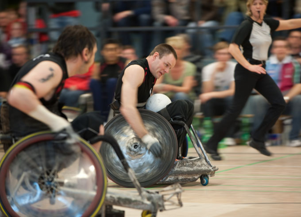 Bernd Best Turnier: zwei Sportler bewegen sich rasant im Rollstuhl voran, um den Ball ins gegnerische Tor zu befördern. Die Linienrichterin läuft im Hintergrund mit.