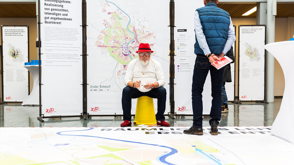 Das Foto zeigt eine teilnehmende Person des zweiten Zukunftsforums an einer Arbeitsstation. Eine zweite Person informiert sich an den Kartenaushängen.