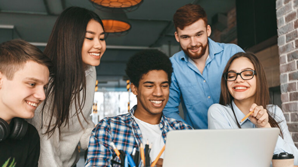 5 junge Menschen sitzen lachelnd vor einem Laptop, Link auf Die Freiwillige Versicherung - Stadt Köln