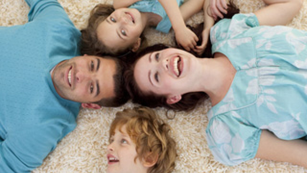 Mutter, Vater und zwei Kinder liegen gemeinsam auf einem Teppich und schauen lachend nach oben.
