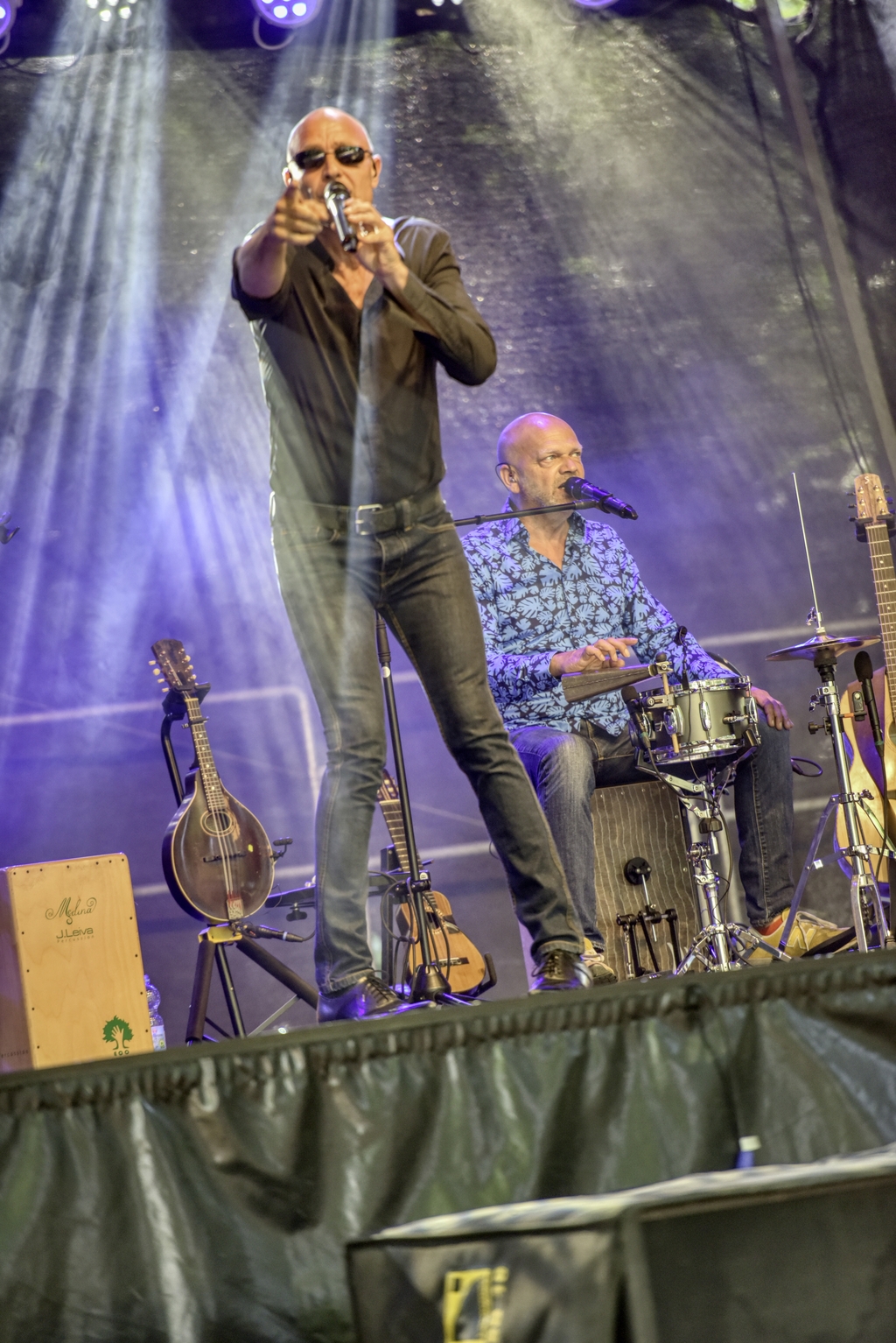 Die beiden Künstler sitzen und stehen auf der Bühne und im Hintergrund stehen verschiedene Instrumente.