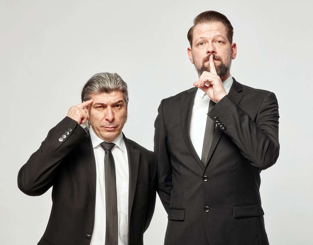 Onkel Fisch - zwei Männer im Anzug, der eine zeigt den Vogel, der andere macht das Leisezeichen mit dem Finger am Mund