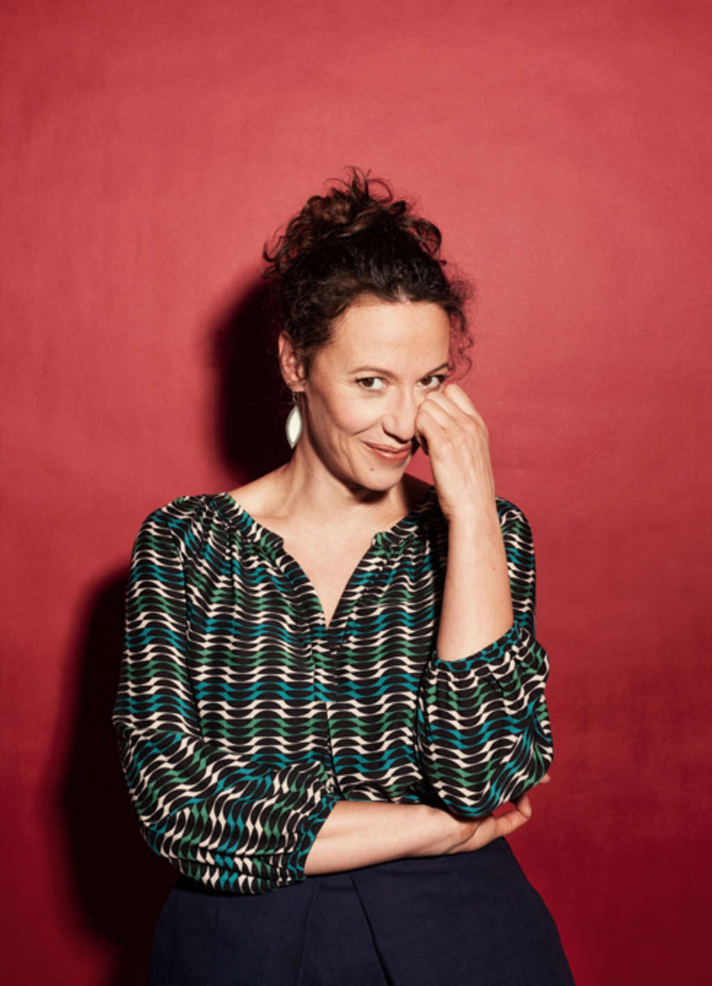 Alice Köfer steht vor einer roten Wand, hält die linke Hand am Gesicht und schaut verschmitzt in die Kamera