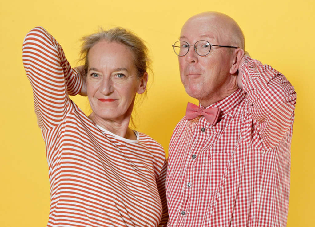 Frau mit geringeltem Shirt und Mann mit kariertem Hemd vor gelbem Hintergrund. Beide halten je eine Hand an den Hinterkopf