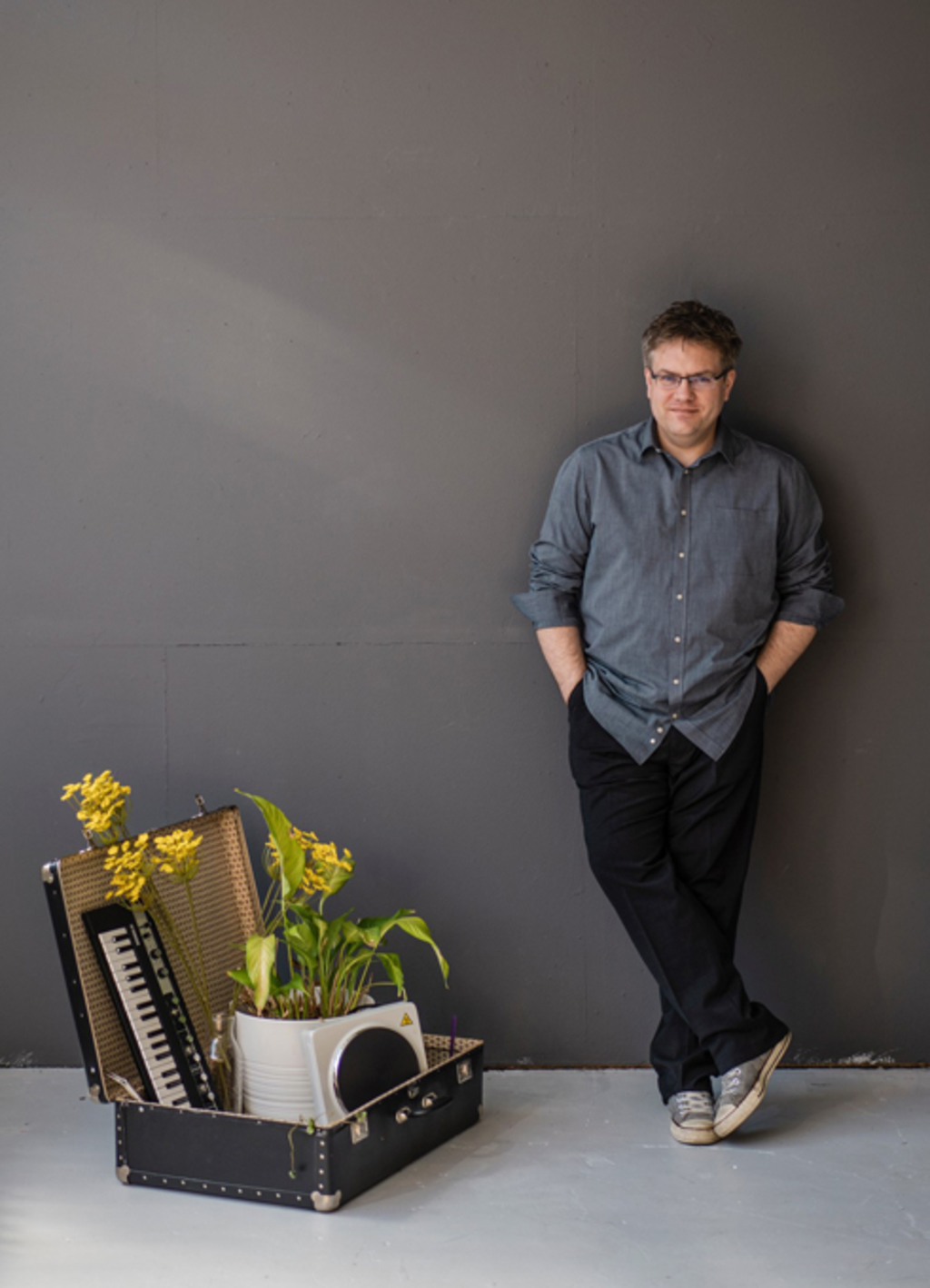 Matthias Reuter lehnt an einer grauen Wand. Neben ihm steht ein geöffneter alter Koffer mit Pflanze, Keyboard und Kochplatte