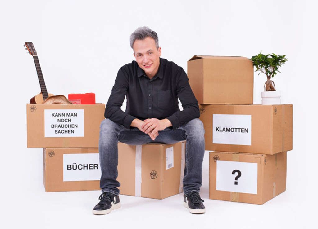 Olaf Bossi sitzt auf einem Umzugkarton, um ihm herum stehen weitere, beschriftete Kartons