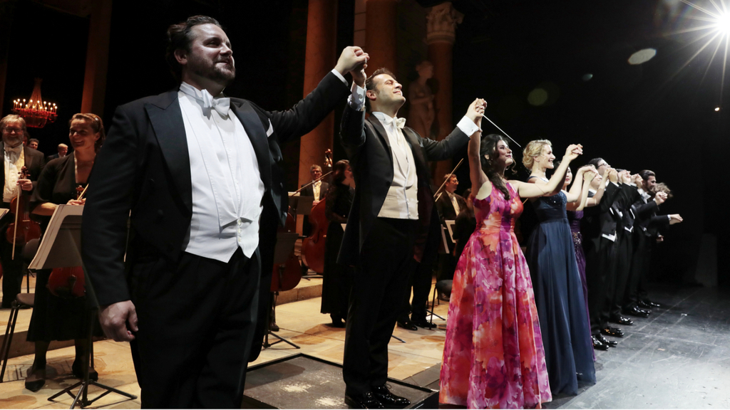 Ein Dirigent und eine Gruppe von Opernsänger*innen verbeugen sich gemeinsam auf der Bühne vor einem Orchester.