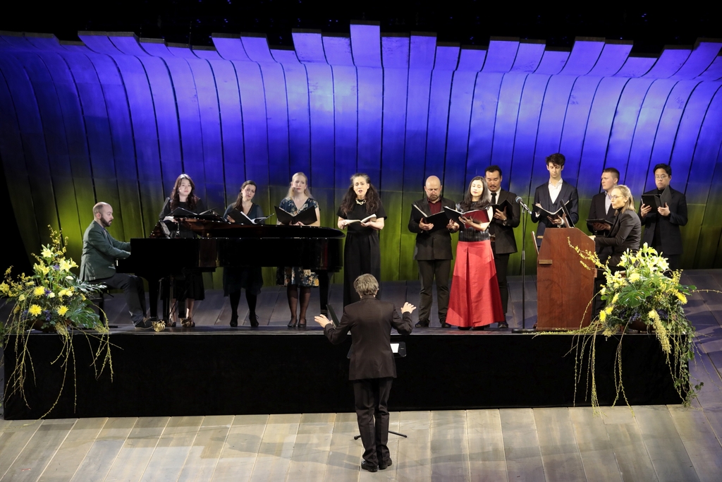 Auf der Bühne singen mehrere Personen, jemand spielt am Flügel, vorne steht ein Dirigent
