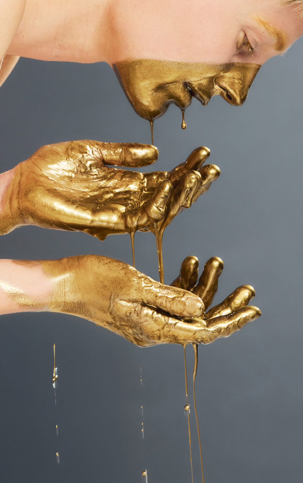 Zwei Hände fangen goldene Farbe auf, die von einem golden bemalten Gesicht tropft.