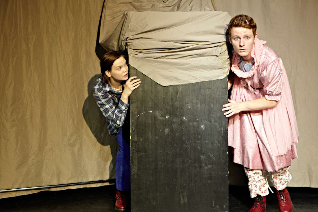 Louisa beziehungsweise Louis in Flanellhemd und Hose und Johann beziehungsweise Johanna in Kleid verstecken sich hinter einem Kasten.