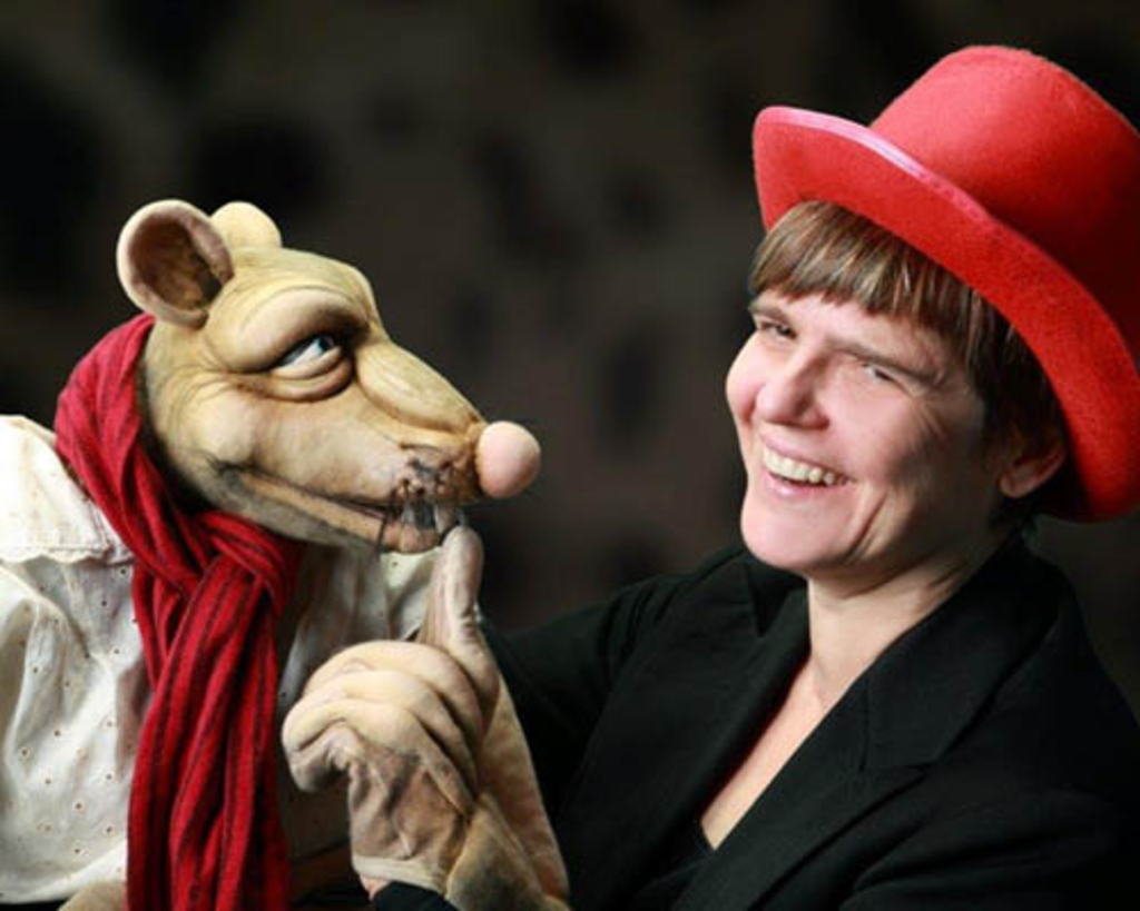 rechts eine Frau mit kurzen Haaren, schwarzem Anzug und rotem Zylinder, sie hält eine Handpuppe - eine Ratte mit weißem Hemd und rotem Schal
