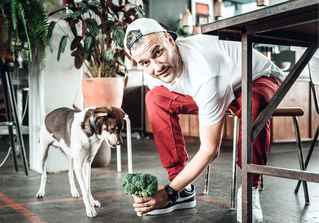 Anton beugt sich unter den Tisch und hält seinem Hund einen Brokkoli hin