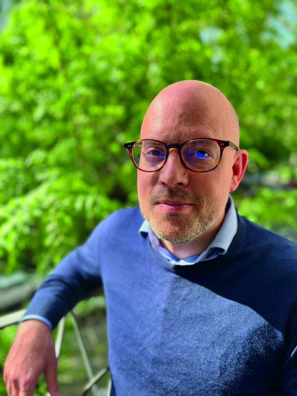 Mann mit Brille sitzt im Garten und schaut lächelnd in die Kamera